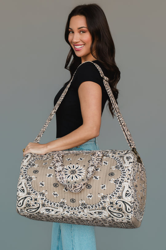 A woman in a black top and jeans smiles while holding the Panache Apparel Collection’s Tan Bandana Duffel, a large patterned beige bag with two handles and a shoulder strap, against a plain background.
