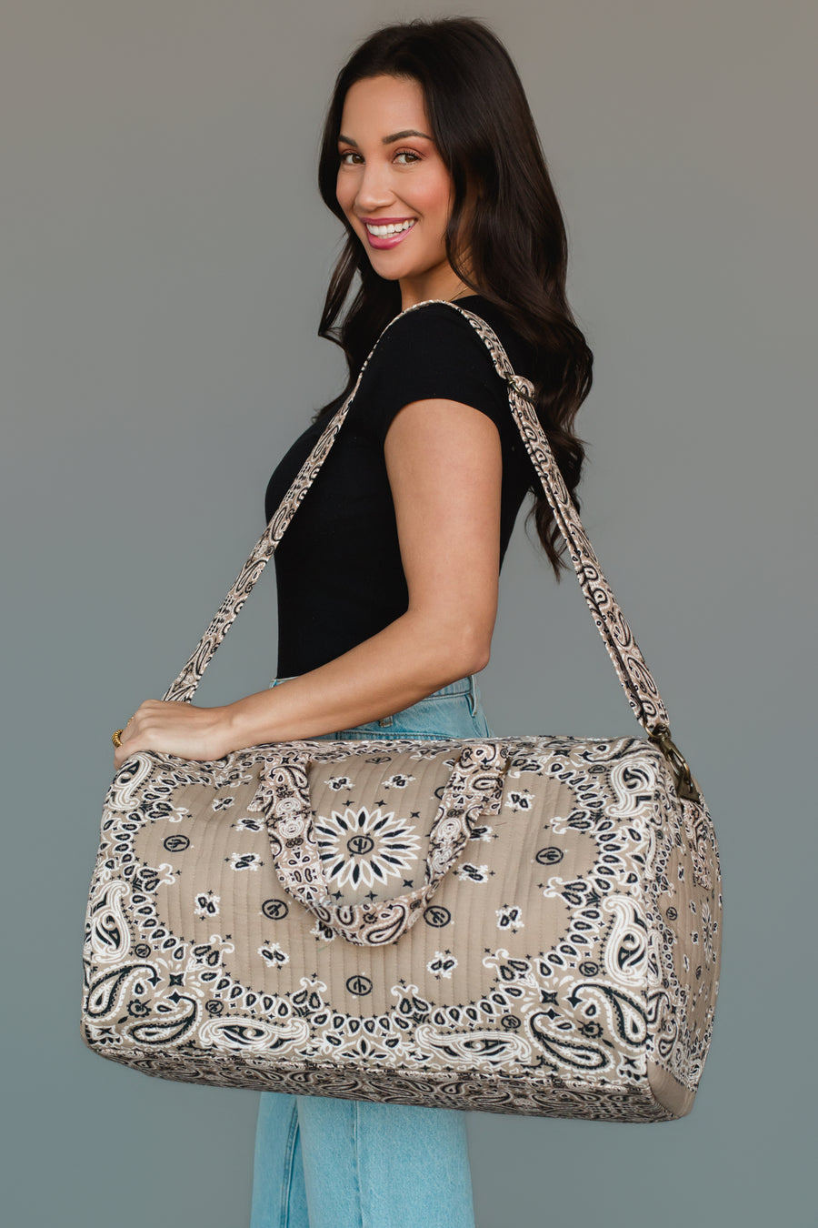 A woman in a black top and jeans smiles while holding the Panache Apparel Collection’s Tan Bandana Duffel, a large patterned beige bag with two handles and a shoulder strap, against a plain background.