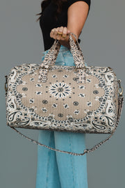 A person wearing a black top and blue jeans holds the Panache Apparel Collection Tan Bandana Duffel, featuring a beige and black bandana pattern, two handles, and a long strap, against a plain background.