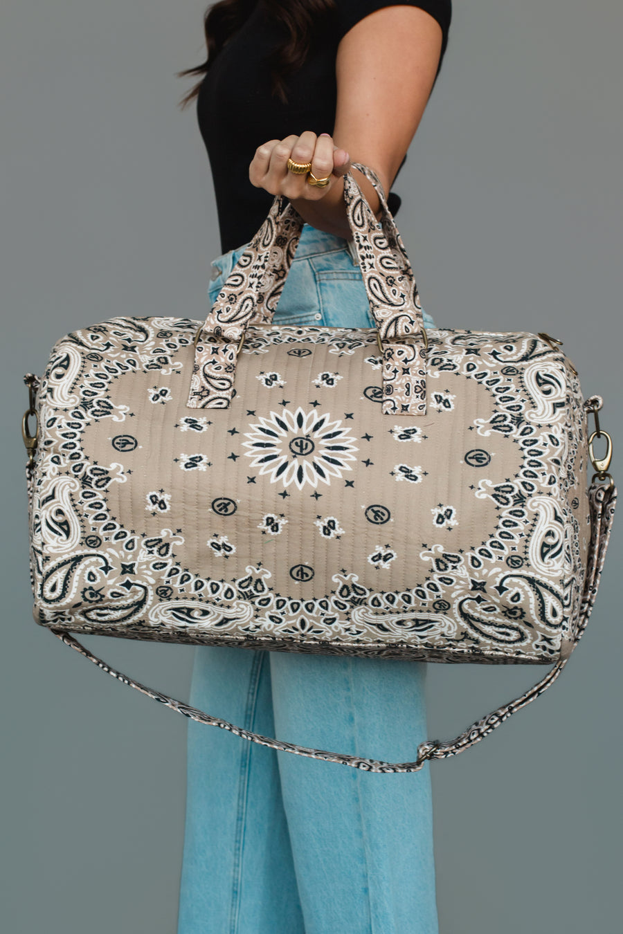A person wearing a black top and blue jeans holds the Panache Apparel Collection Tan Bandana Duffel, featuring a beige and black bandana pattern, two handles, and a long strap, against a plain background.