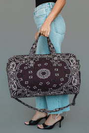 A person in blue jeans and black heels holds the Panache Apparel Collection Brown Bandana Duffel, featuring a white paisley design and shoulder strap, against a plain gray background.
