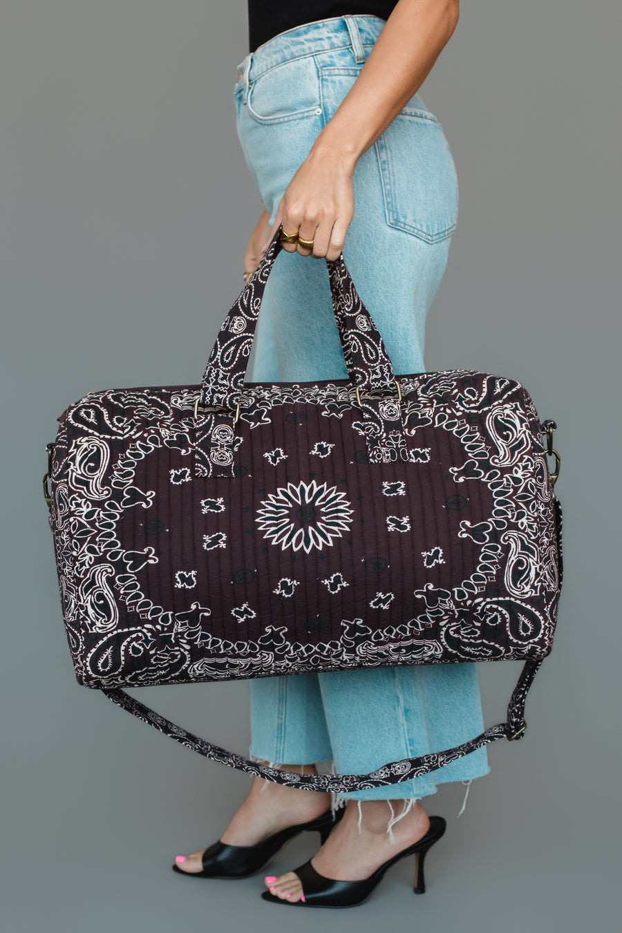 A person in blue jeans and black heels holds the Panache Apparel Collection Brown Bandana Duffel, featuring a white paisley design and shoulder strap, against a plain gray background.