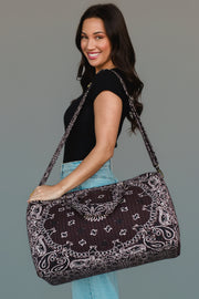 A woman in a black shirt and light blue jeans smiles while holding the Panache Apparel Collection Brown Bandana Duffel, which features both shoulder and hand straps.
