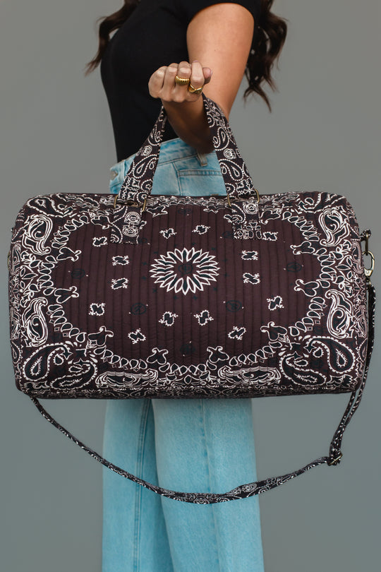 A person in a black shirt and blue jeans holds the Panache Apparel Collection Brown Bandana Duffel, featuring a brown and white paisley design with a matching shoulder strap, against a gray background.