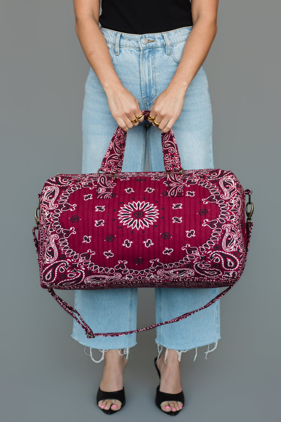 A person wearing a black top, light blue jeans, and black heels holds the Burgundy Bandana Duffel by Panache Apparel Collection with a shoulder strap against a plain gray background.