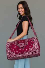 A smiling woman with long dark hair wears a black shirt and jeans, holding the Panache Apparel Collection Burgundy Bandana Duffel over her shoulder, standing against a plain gray background.