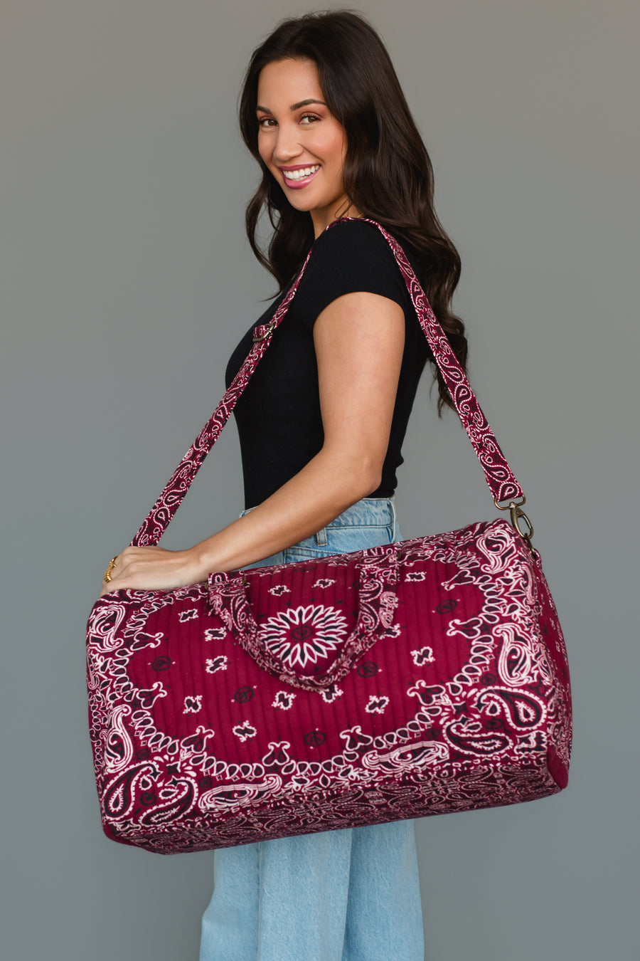 A smiling woman with long dark hair wears a black shirt and jeans, holding the Panache Apparel Collection Burgundy Bandana Duffel over her shoulder, standing against a plain gray background.