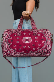 A person wearing a black top and blue jeans holds the Panache Apparel Collection Burgundy Bandana Duffel, which features handles and a shoulder strap, against a plain gray background.