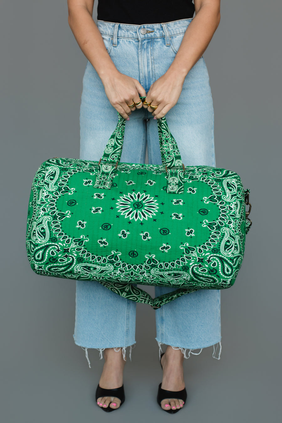 A person in a black top and light blue jeans holds the Panache Apparel Collection Green Bandana Duffel with both hands against a gray background.