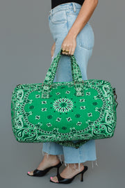 A person dressed in light blue jeans and black heels holds the Panache Apparel Collection Green Bandana Duffel with both hands against a plain gray background.