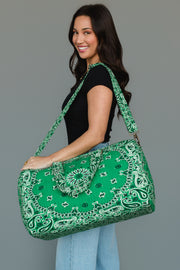 A woman in a black shirt and light blue jeans smiles as she carries the Panache Apparel Collection’s Green Bandana Duffel over her shoulder, standing against a plain gray background.