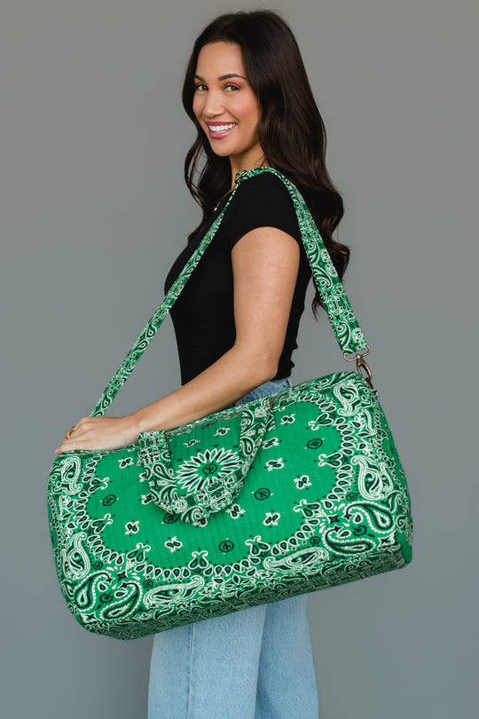 A woman in a black shirt and light blue jeans smiles as she carries the Panache Apparel Collection’s Green Bandana Duffel over her shoulder, standing against a plain gray background.