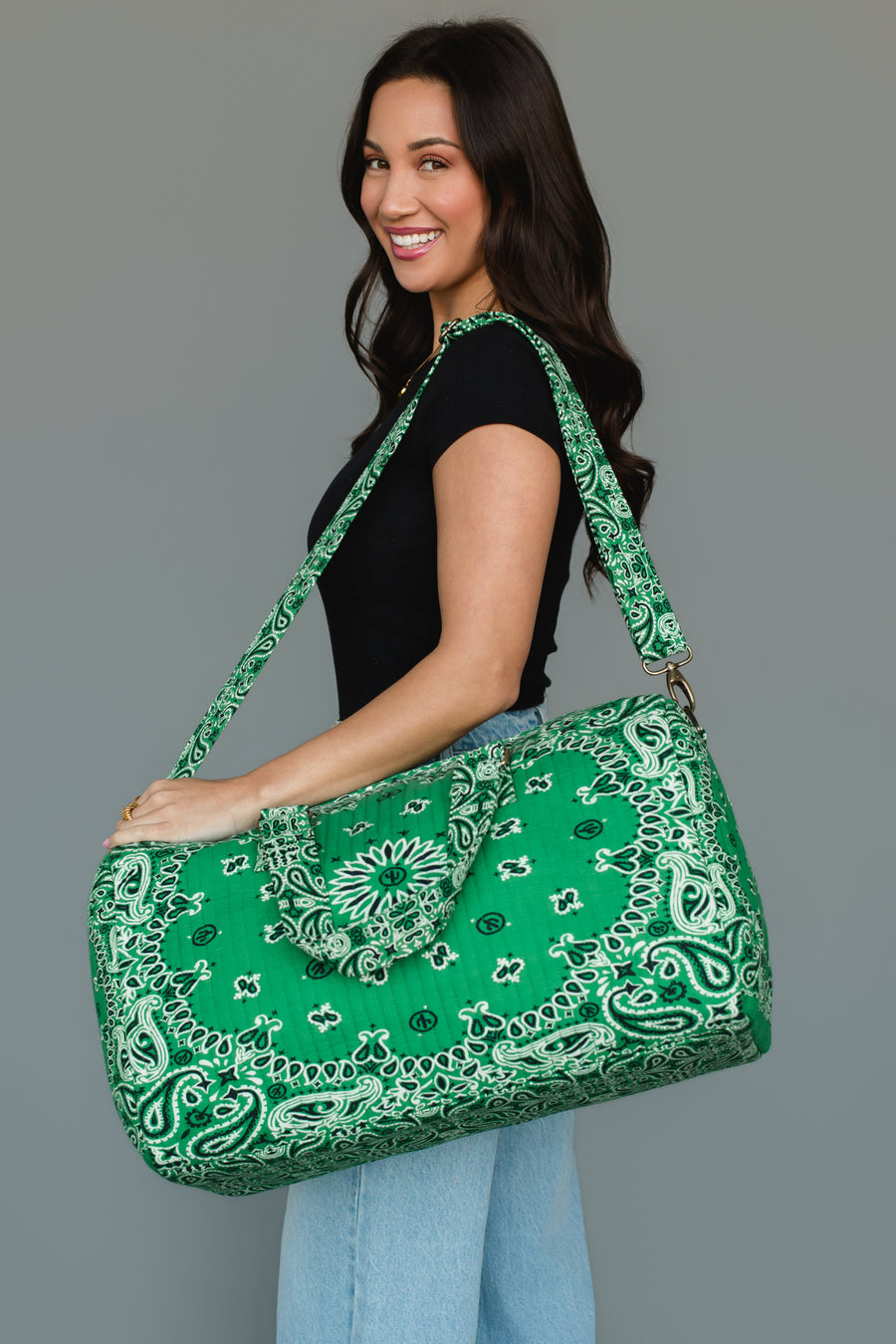 A woman in a black shirt and light blue jeans smiles as she carries the Panache Apparel Collection’s Green Bandana Duffel over her shoulder, standing against a plain gray background.