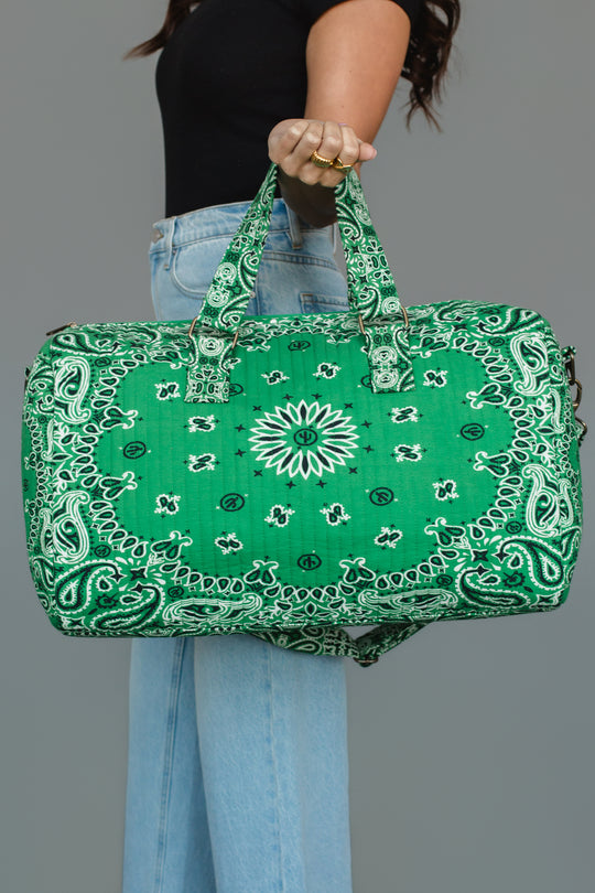 A person in a black top and blue jeans holds the Panache Apparel Collection Green Bandana Duffel, featuring a white paisley bandana pattern, against a plain gray background.