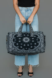 A person in light blue jeans and a black top holds the Black Bandana Duffel by Panache Apparel Collection, featuring a bold black and white paisley pattern. The individual stands against a gray background, with their head out of frame.