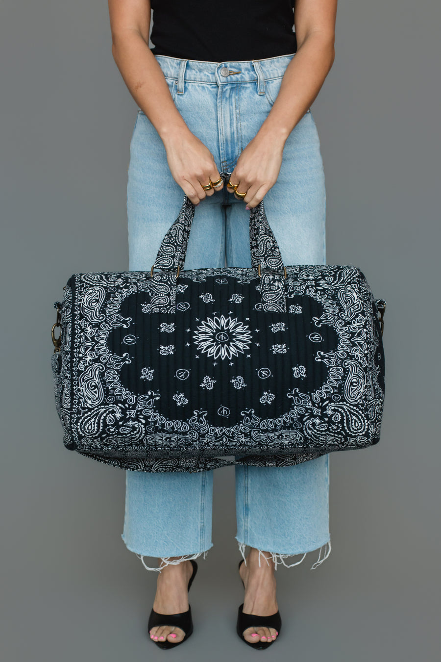 A person in light blue jeans and a black top holds the Black Bandana Duffel by Panache Apparel Collection, featuring a bold black and white paisley pattern. The individual stands against a gray background, with their head out of frame.