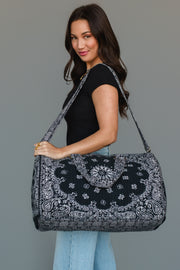 A woman with long dark hair, dressed in a black top and light jeans, stands against a plain gray background while carrying the Panache Apparel Collection Black Bandana Duffel, a large black duffel bag featuring a white paisley pattern, over her shoulder.