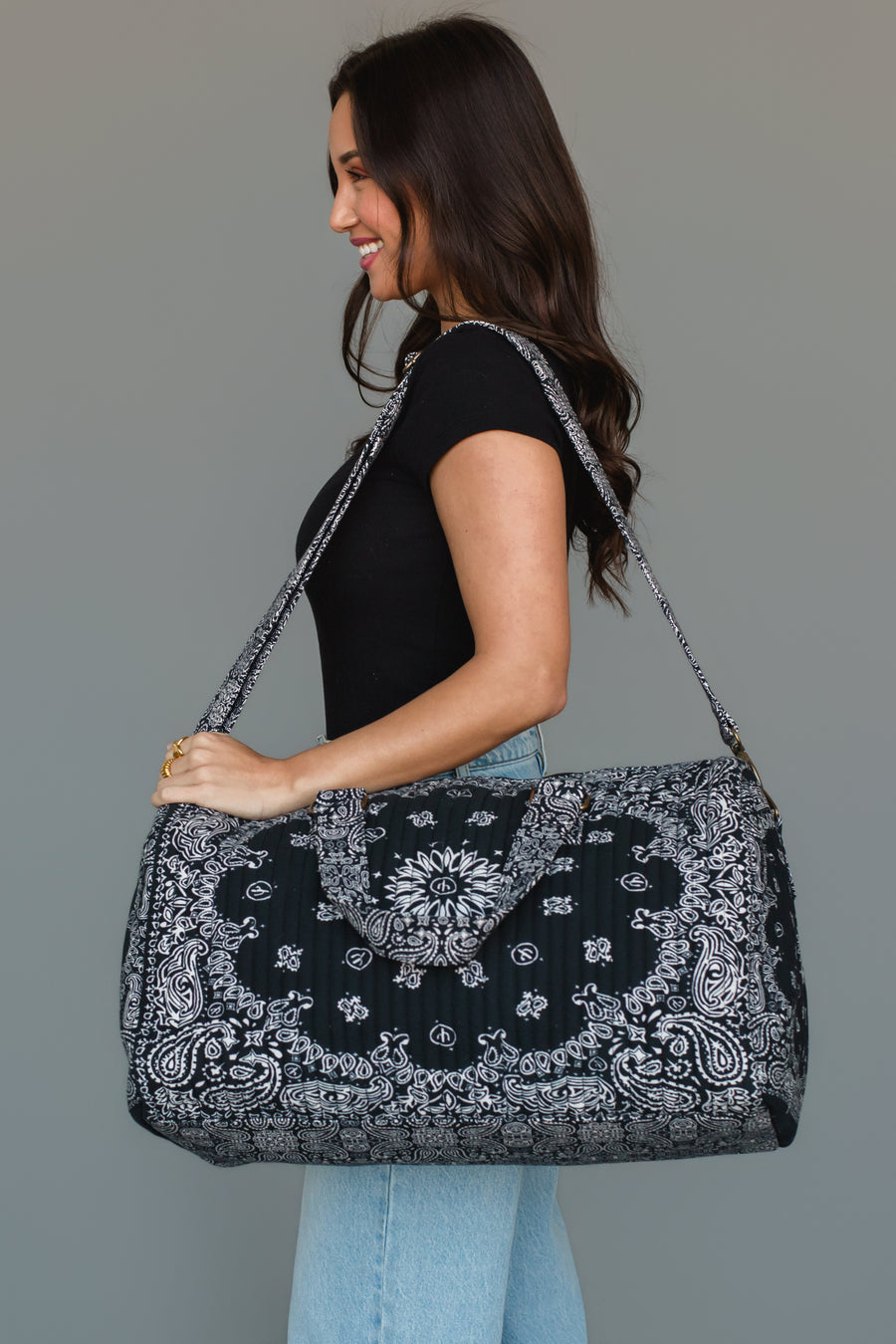A woman wearing a black shirt and light blue jeans stands in profile against a plain gray background, holding the Panache Apparel Collection Black Bandana Duffel featuring a large black and white paisley pattern.