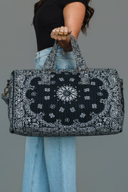 A person in a black shirt and blue jeans holds the Panache Apparel Collection’s Black Bandana Duffel with both hands against a plain gray background.