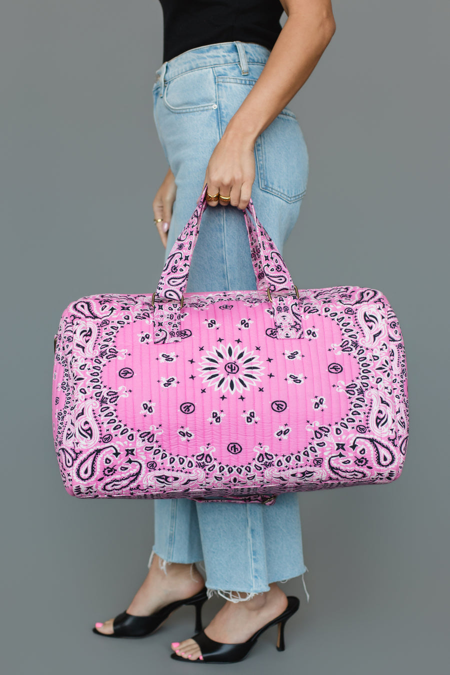 A person in blue jeans and black heels holds the Panache Apparel Collection Pink Bandana Duffel against a plain gray background.