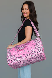 A woman in a black top and light blue jeans smiles while holding the Pink Bandana Duffel by Panache Apparel Collection, featuring a large pink quilted design with a paisley pattern, over her shoulder against a plain gray background.