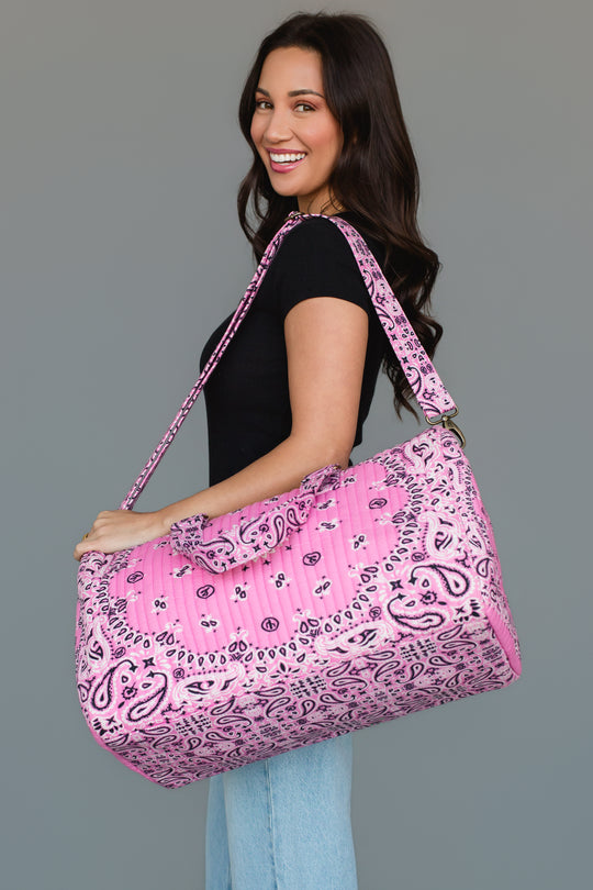 A woman in a black top and light blue jeans smiles while holding the Pink Bandana Duffel by Panache Apparel Collection, featuring a large pink quilted design with a paisley pattern, over her shoulder against a plain gray background.