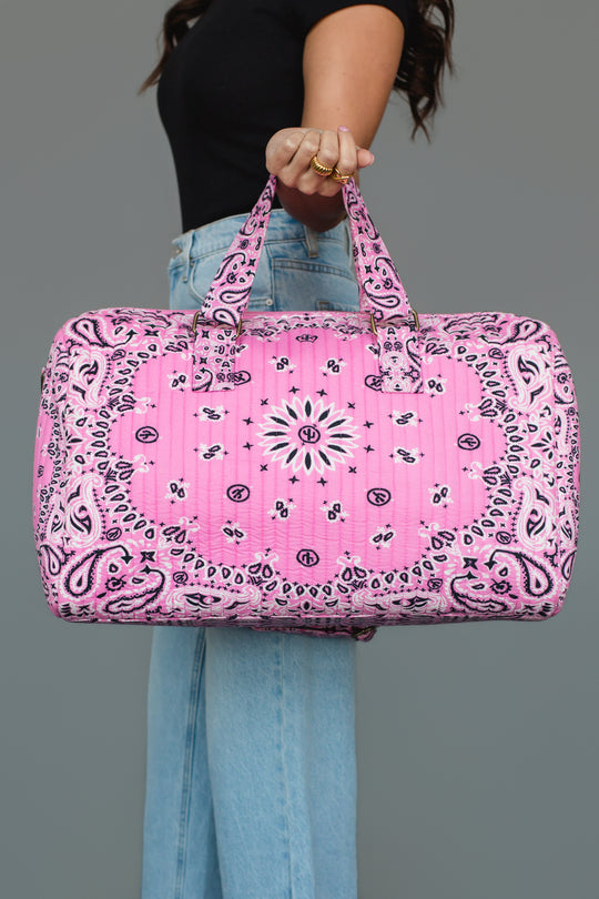 A person in a black top and blue jeans holds the Pink Bandana Duffel by Panache Apparel Collection by its handles against a gray background.