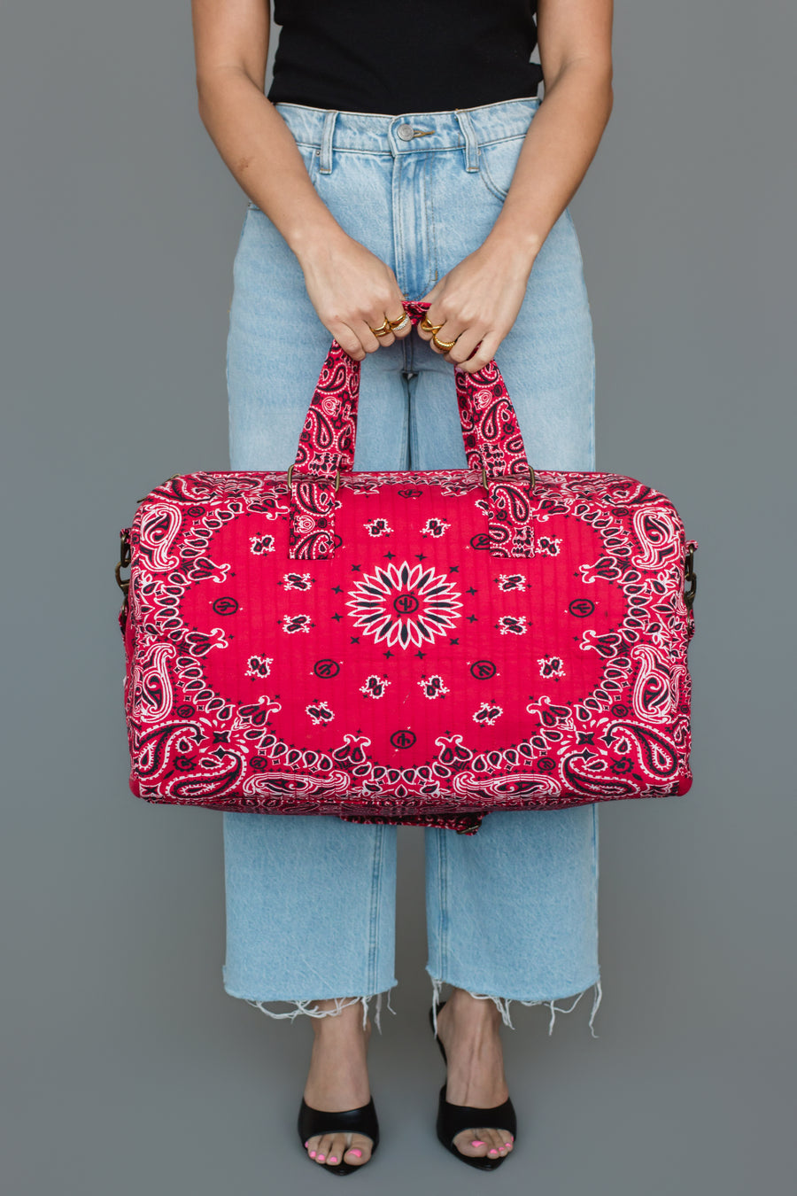 Wearing light blue jeans and a black top, an individual holds the Panache Apparel Collection’s Red Bandana Duffel with both hands. Set against a gray background, only the person’s torso, legs, and feet in black sandals are visible.