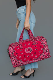 A woman in jeans and a black top stands against a gray background, wearing black heels and holding the Panache Apparel Collection’s Red Bandana Duffel.