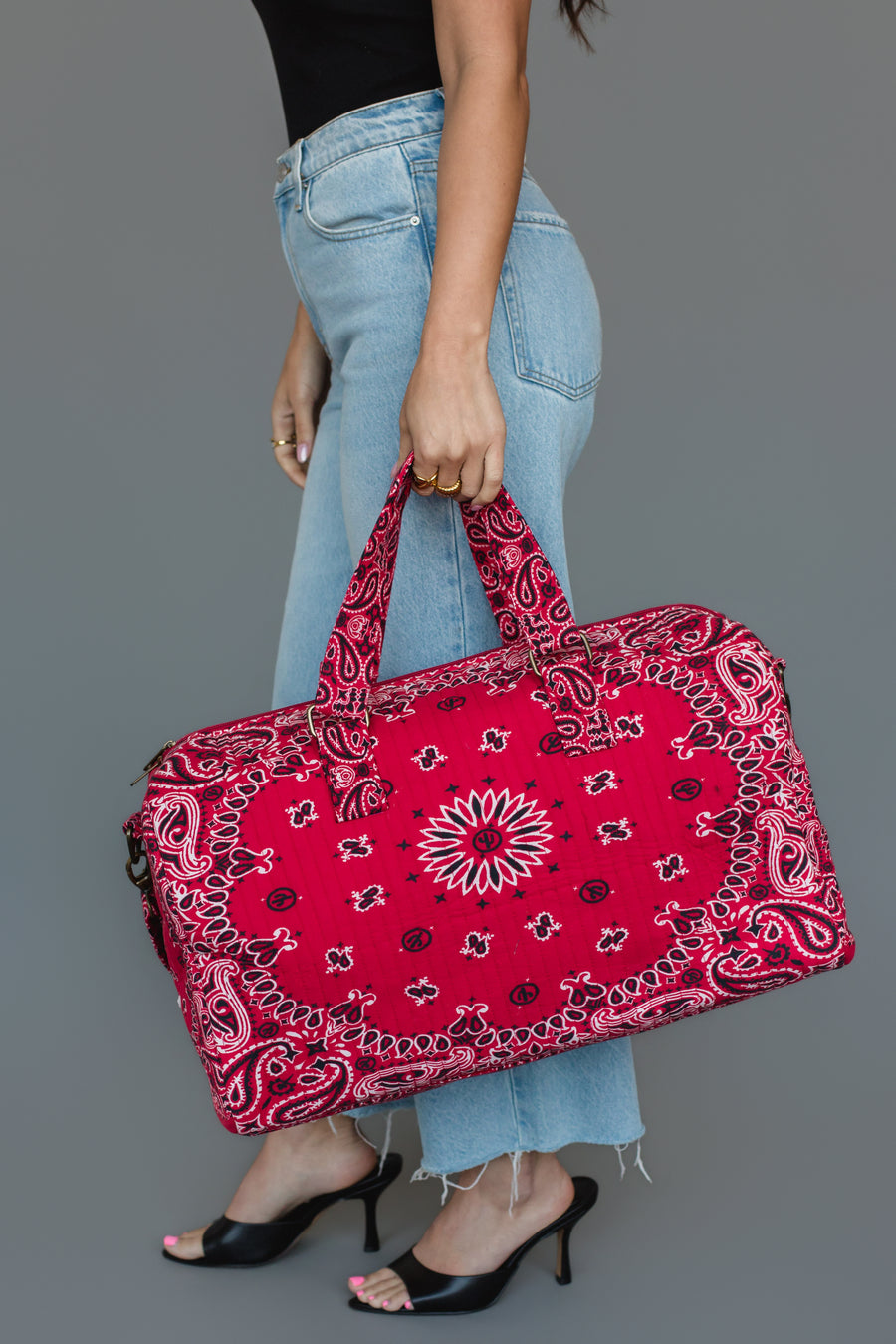A woman in jeans and a black top stands against a gray background, wearing black heels and holding the Panache Apparel Collection’s Red Bandana Duffel.