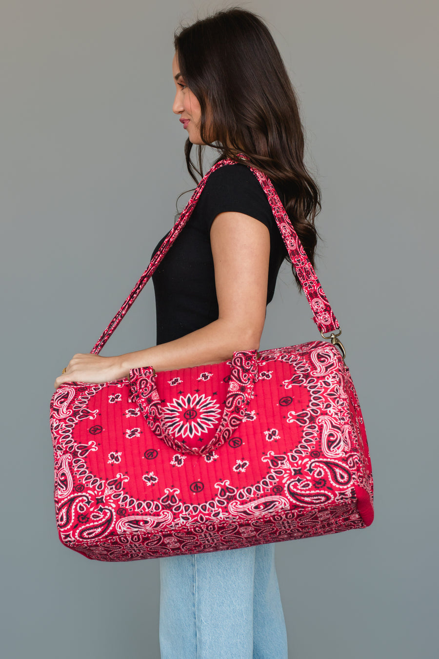 A woman in a black shirt and light blue jeans holds the Panache Apparel Collection Red Bandana Duffel, featuring a bold red paisley pattern and matching shoulder strap, against a plain gray background.