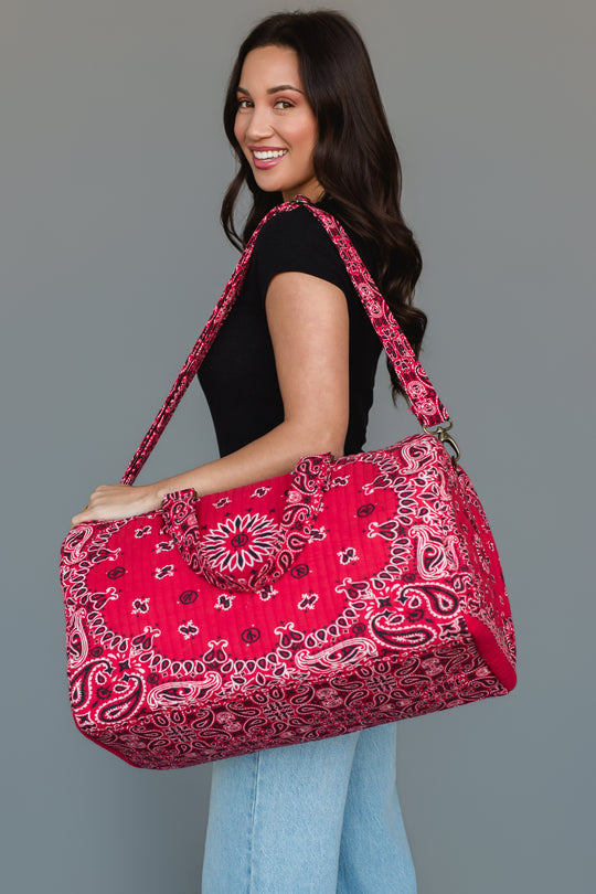 A woman smiles and poses in a black shirt and light blue jeans, carrying the Panache Apparel Collection’s Red Bandana Duffel, a large, bright red patterned bag with a shoulder strap, against a plain gray background.