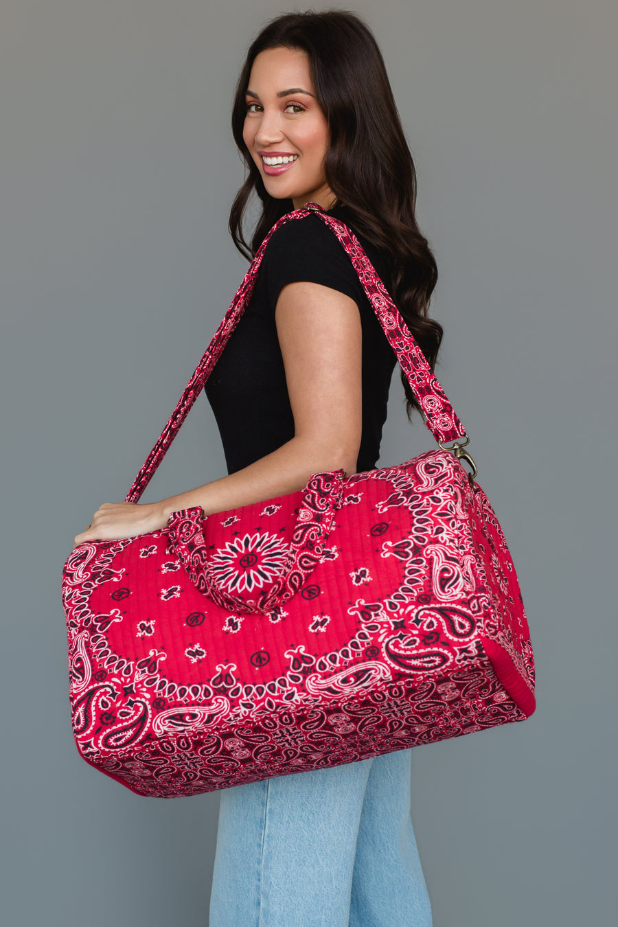 A woman smiles and poses in a black shirt and light blue jeans, carrying the Panache Apparel Collection’s Red Bandana Duffel, a large, bright red patterned bag with a shoulder strap, against a plain gray background.
