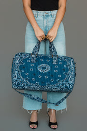 A person in light blue jeans and a black top holds the Panache Apparel Collection Dark Blue Bandana Duffel with both hands. They wear open-toed shoes and stand against a plain gray background.