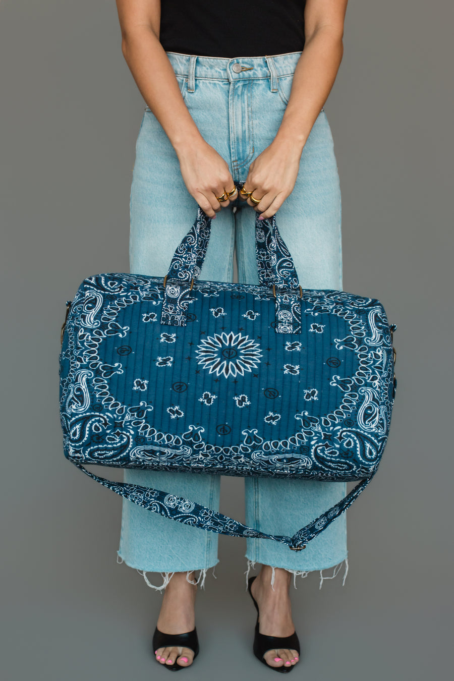 A person in light blue jeans and a black top holds the Panache Apparel Collection Dark Blue Bandana Duffel with both hands. They wear open-toed shoes and stand against a plain gray background.