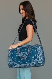 A smiling woman in a black shirt and light blue jeans holds the Panache Apparel Collection’s Dark Blue Bandana Duffel, featuring a bold blue and white pattern and a shoulder strap, against a plain gray background.