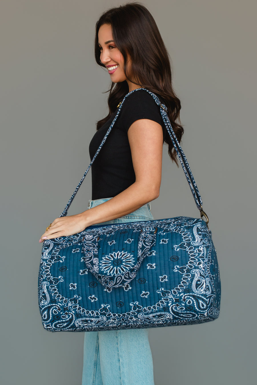 A smiling woman in a black shirt and light blue jeans holds the Panache Apparel Collection’s Dark Blue Bandana Duffel, featuring a bold blue and white pattern and a shoulder strap, against a plain gray background.