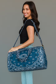A woman smiles as she carries the Panache Apparel Collection Dark Blue Bandana Duffel over her shoulder, wearing a black shirt and light blue jeans against a plain gray background.