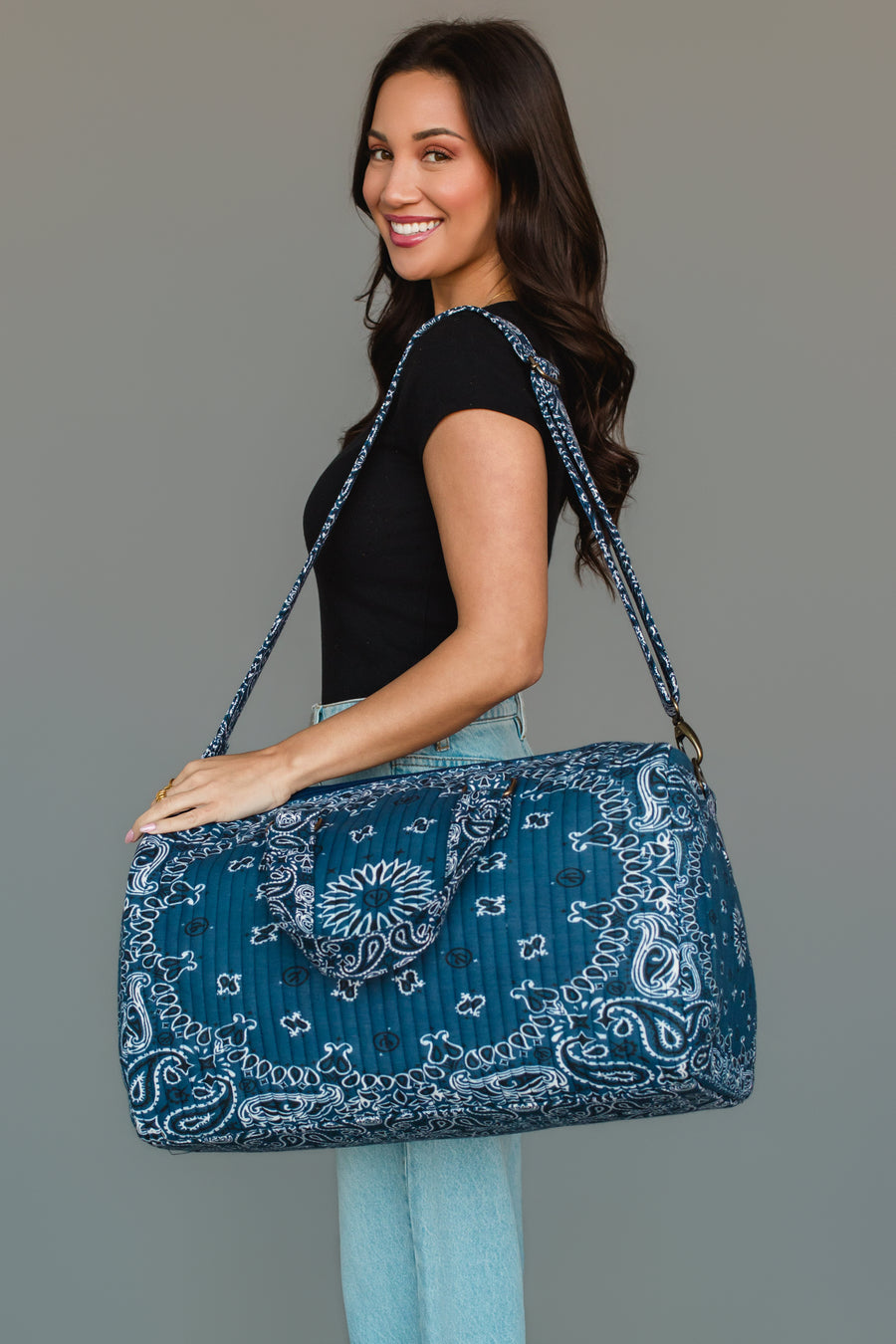 A woman smiles as she carries the Panache Apparel Collection Dark Blue Bandana Duffel over her shoulder, wearing a black shirt and light blue jeans against a plain gray background.