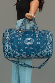 A person in jeans and a black top holds the Panache Apparel Collection Dark Blue Bandana Duffel, which features a quilted design with a white bandana print and both hand and shoulder straps.