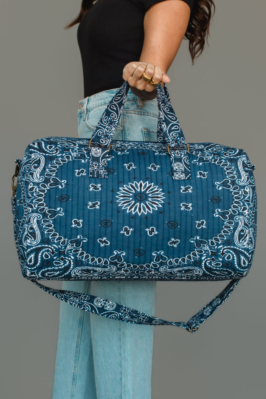A person in jeans and a black top holds the Panache Apparel Collection Dark Blue Bandana Duffel, which features a quilted design with a white bandana print and both hand and shoulder straps.