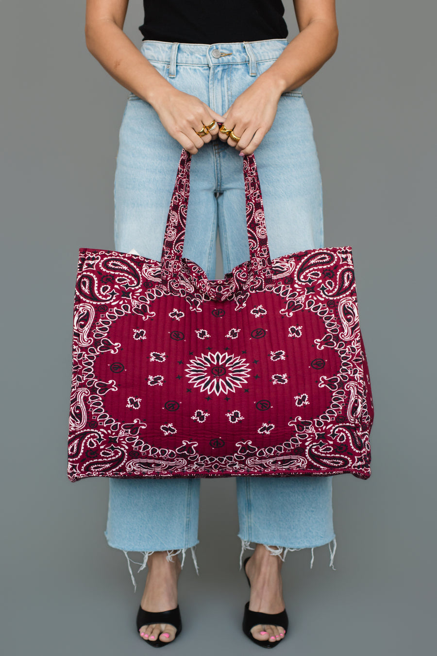 A person wearing light blue jeans and black heels holds the Panache Apparel Collection Burgundy Bandana Tote in front of a gray background.
