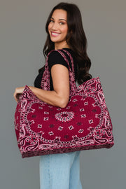 A woman with long dark hair, dressed in a black top and light blue jeans, smiles while carrying the Panache Apparel Collection’s Burgundy Bandana Tote over her shoulder, posing against a plain grey background.