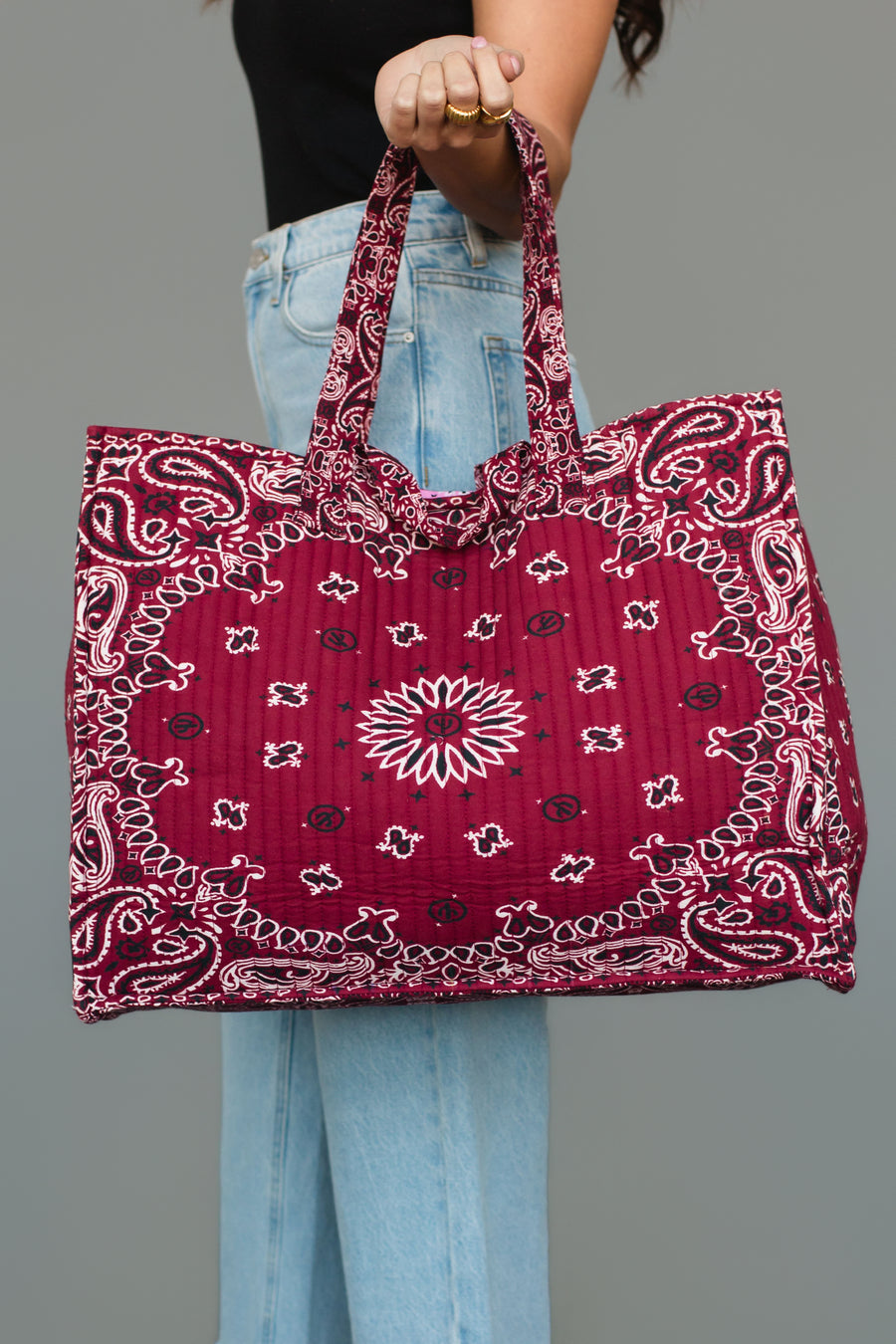 A person wearing light blue jeans and a black top holds the Panache Apparel Collection Burgundy Bandana Tote by its handles against a plain gray background.