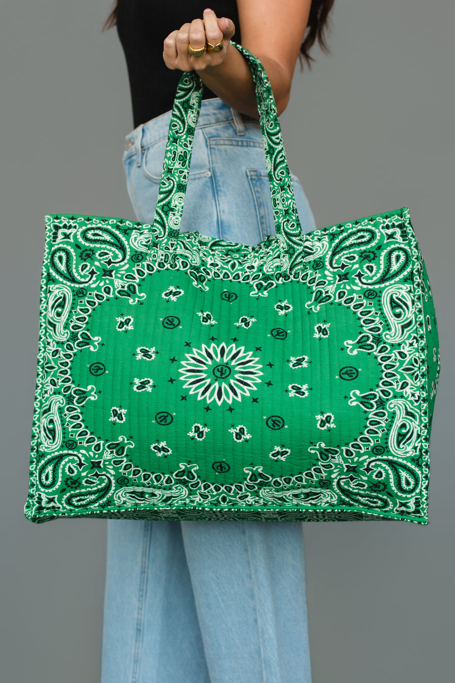 A person wearing light blue jeans and a black top holds the Green Bandana Tote by Panache Apparel Collection, featuring a large green design with a white bandana print, against a plain gray background.