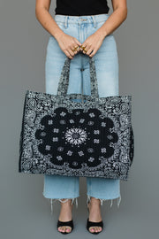 A person dressed in light blue jeans and a black top holds the Panache Apparel Collection’s Black Bandana Tote, a spacious tote bag featuring a bold black and white paisley pattern and two sturdy handles. They are standing against a plain gray background.