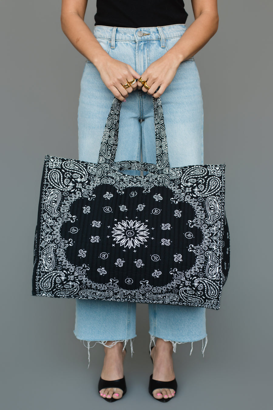 A person dressed in light blue jeans and a black top holds the Panache Apparel Collection’s Black Bandana Tote, a spacious tote bag featuring a bold black and white paisley pattern and two sturdy handles. They are standing against a plain gray background.