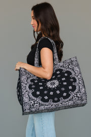 A woman with long brown hair, dressed in a black top and light blue jeans, carries the Panache Apparel Collection Black Bandana Tote, an oversized bag featuring a black and white paisley pattern, over her shoulder, standing against a neutral gray background.