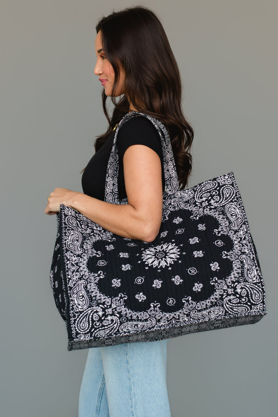 A woman with long brown hair, dressed in a black top and light blue jeans, carries the Panache Apparel Collection Black Bandana Tote, an oversized bag featuring a black and white paisley pattern, over her shoulder, standing against a neutral gray background.
