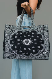 A person wearing a black top and light blue jeans holds the Panache Apparel Collection Black Bandana Tote, a large rectangular bag featuring a black and white paisley bandana print and two handles, against a gray background.